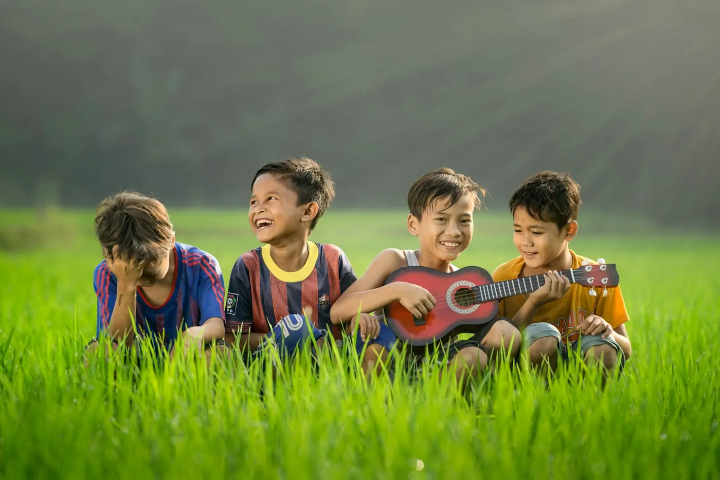 Empat anak laki-laki tertawa sambil bermain ukulele di sawah